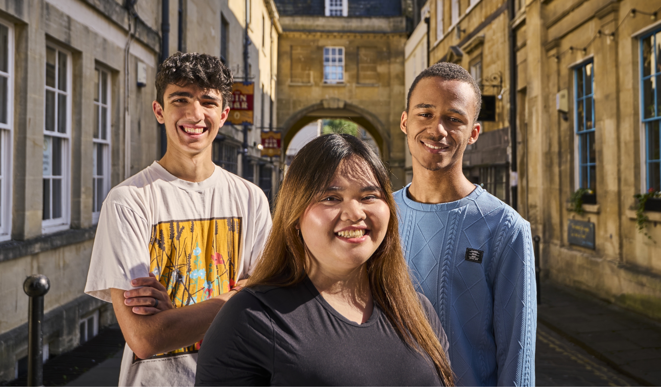 Three people stand smiling in front of an archway and historic looking buildings.