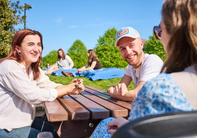 Students sat together outside at a picnic bench, with others in the background sat on large outdoor cushions.