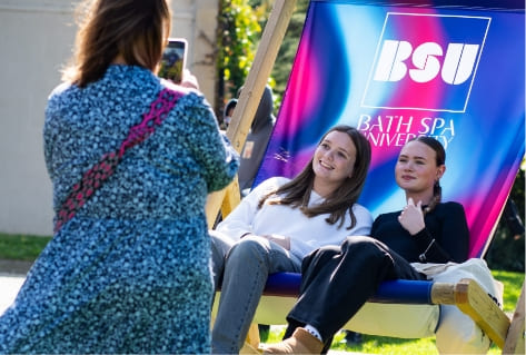 Two people sit on a large BSU branded deck chair, another person is taking a photo of them on their mobile.