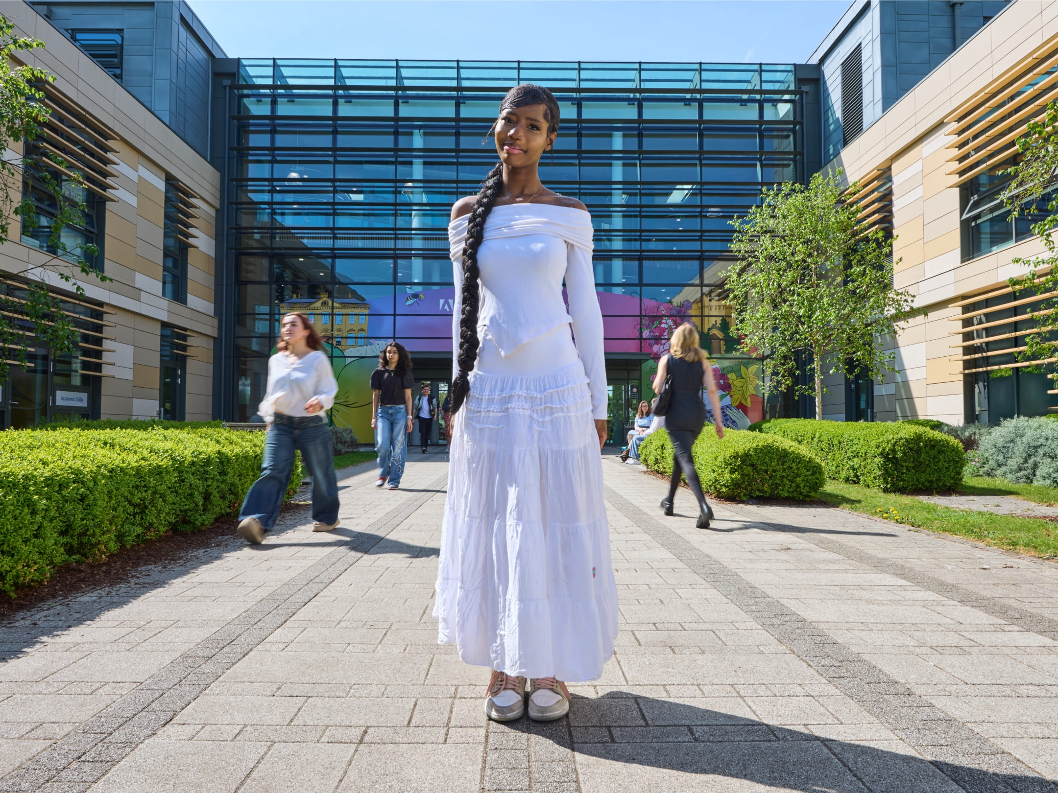 A student stood centrally in a walkway, with a large glass fronted building behind. Other students walk by casually.