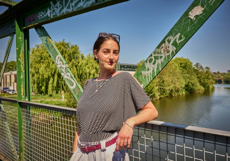 A person standing casually on a graffiti-covered metal bridge overlooking a calm river and green trees on a sunny day.
