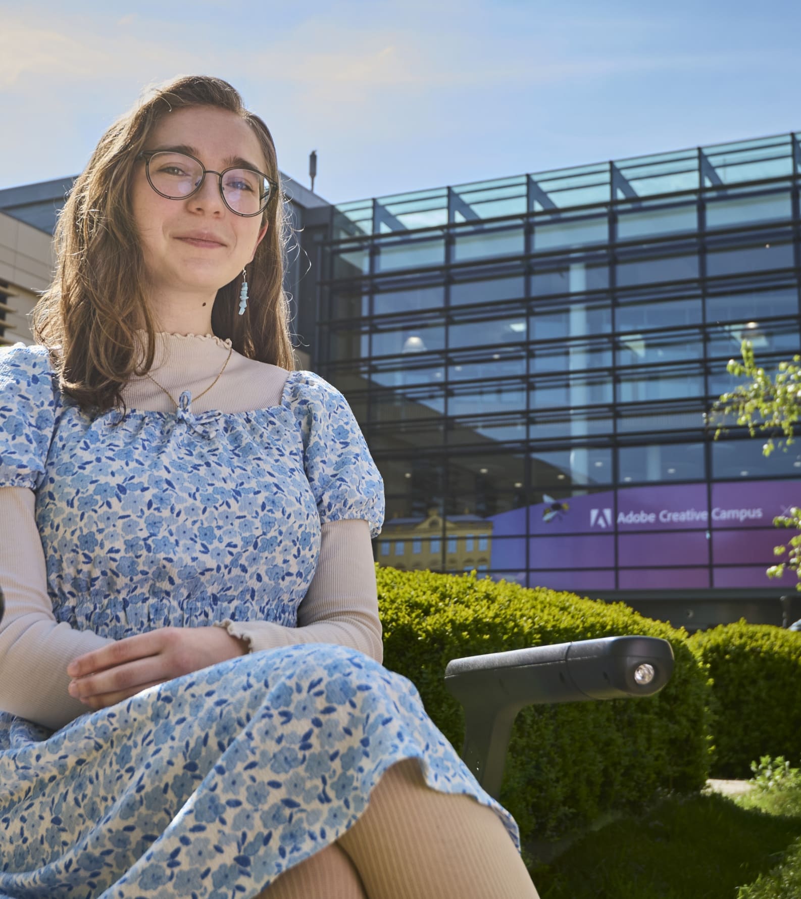 A person sat on a wheelchair in front of a modern, glass fronted building on a sunny day.
