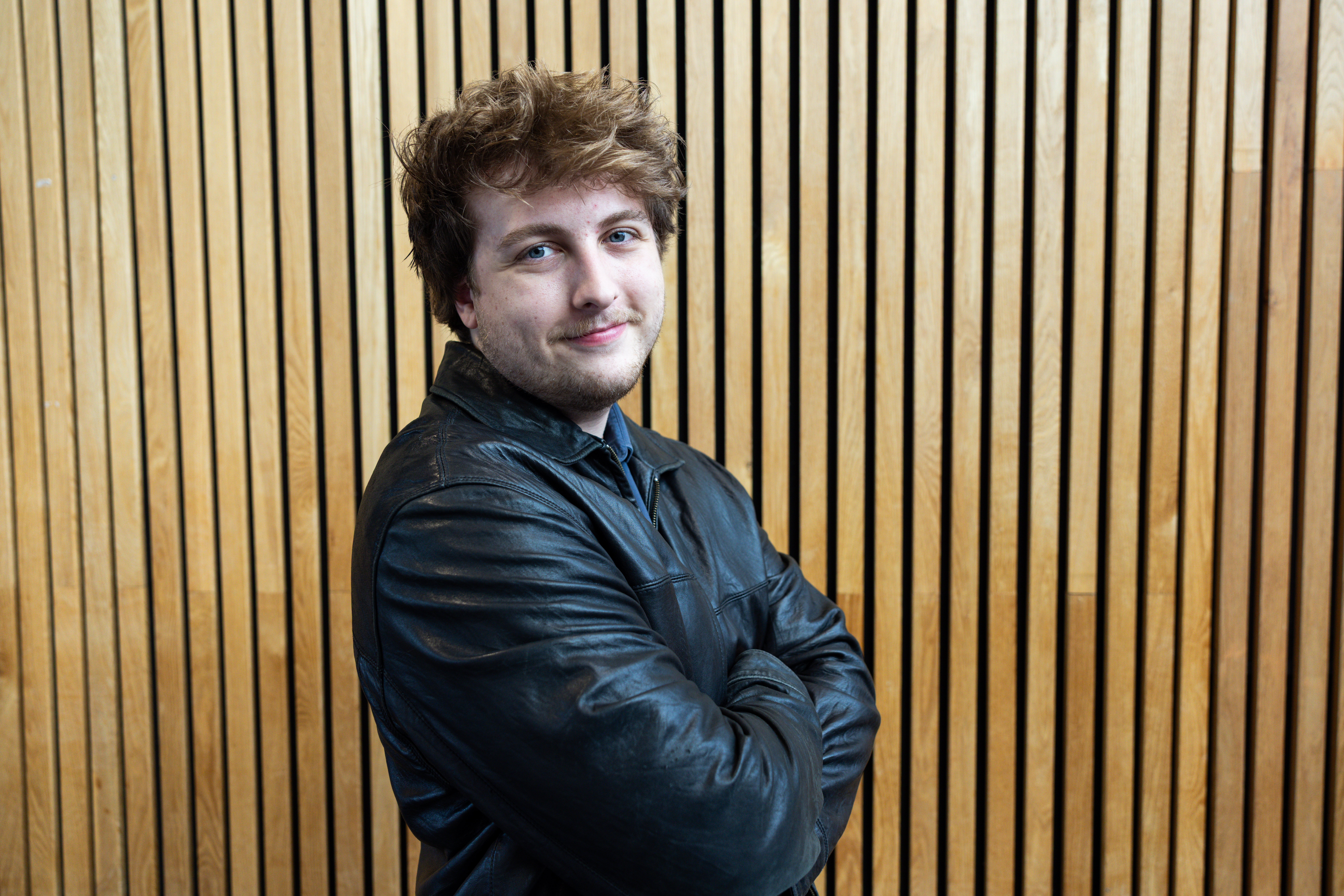A student is smiling towards the camera but stood off centre. Behind the student is a slated wall.