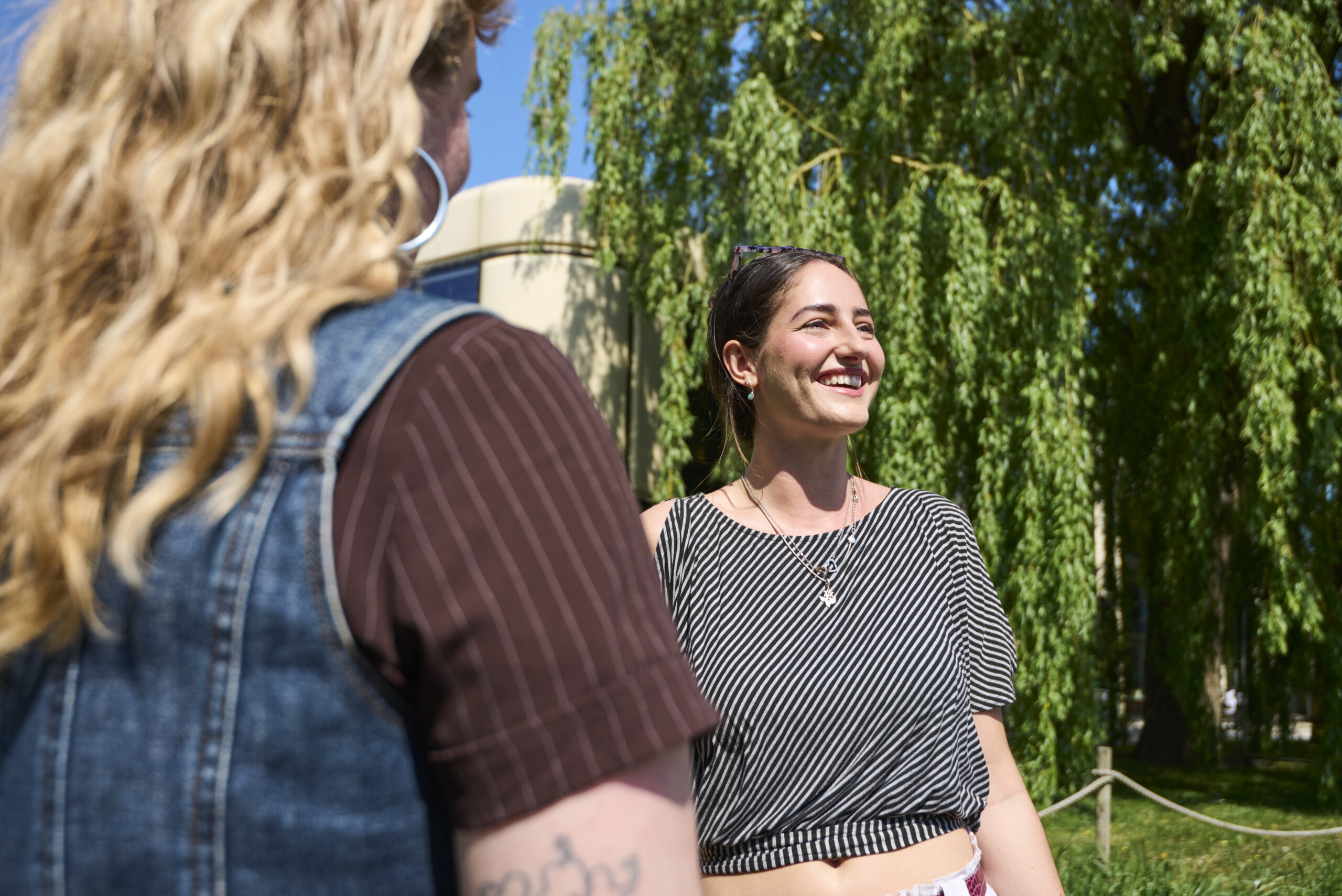 One student smiling towards something off camera and another students back can be seen closer to the camera. It's a sunny day and there is a large leafy tree behind the student.