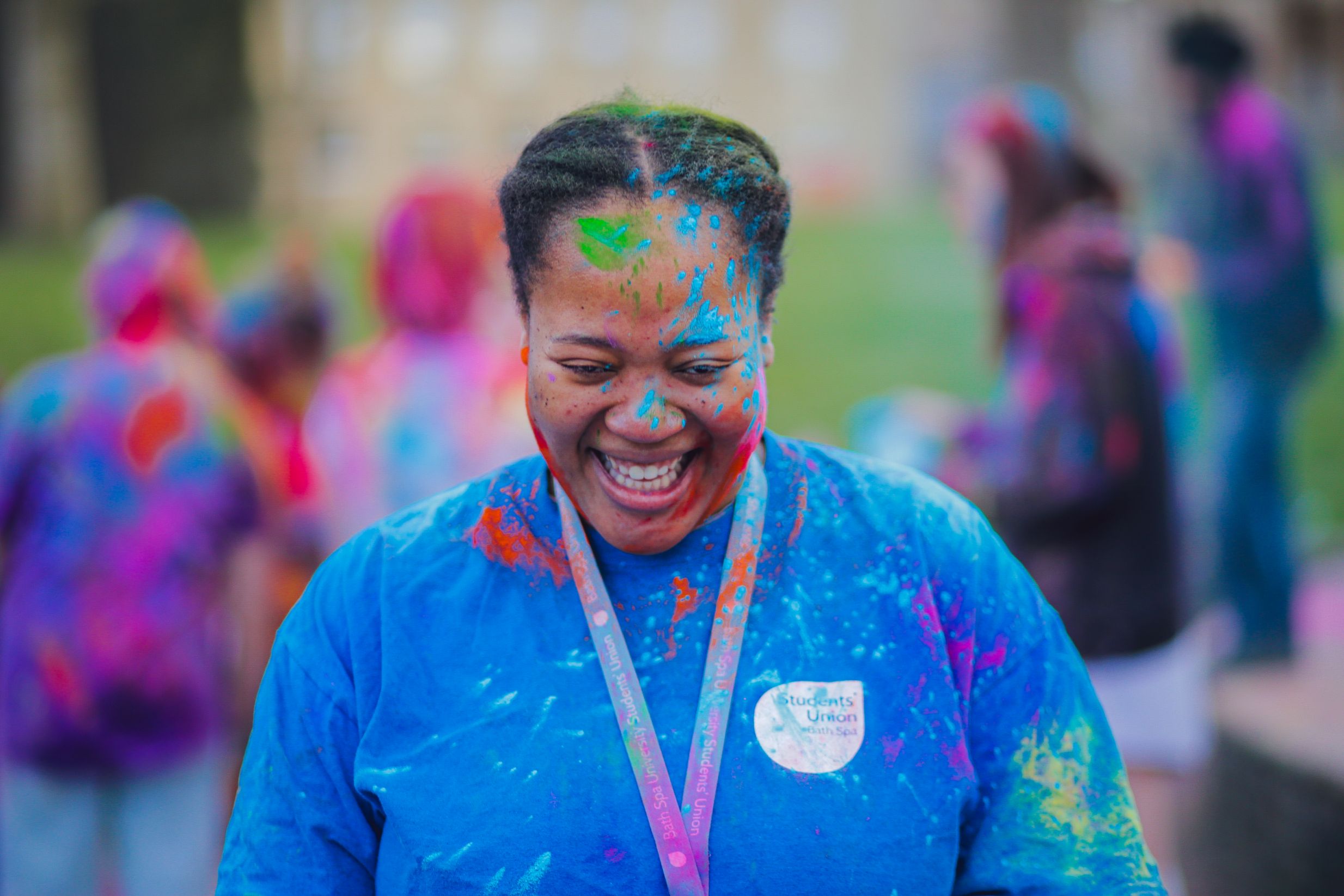 A person close to the camera is covered in UV paint and is wearing a Bath Spa Students Union blue t-shirt. There are blurred people in the background.