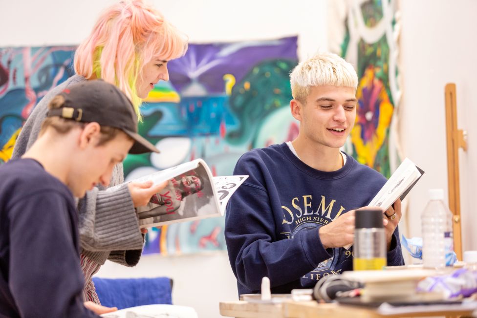 Three students looking through books and magazines in front of a colourful background.